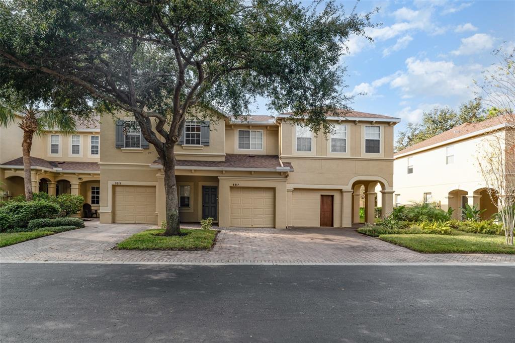 A two-story tan stucco townhouse with two garages and a paved driveway under a tree — 9317 STONE RIVER PLACE, RIVERVIEW, FL