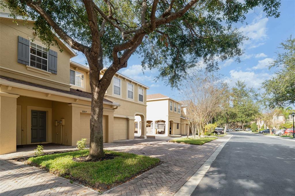A street view of two-story tan townhomes with brick paver driveways and mature trees — 9317 STONE RIVER PLACE, RIVERVIEW, FL