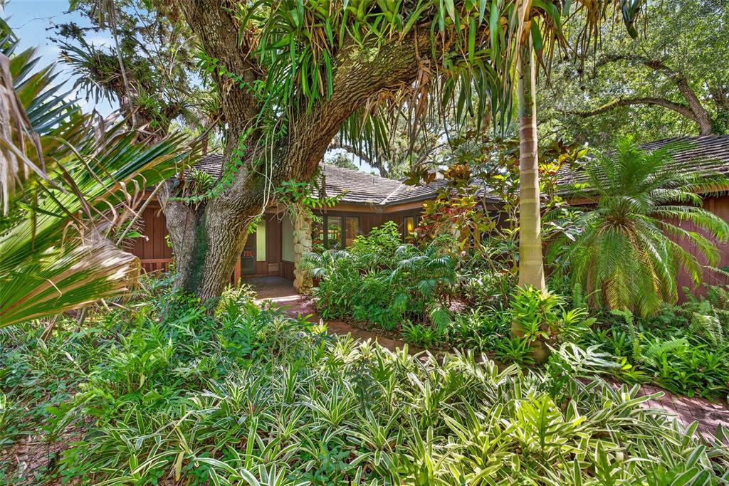 A lush, landscaped front yard leads to the entrance of a wood-sided house under trees — 3460/3480 RIVER RUN LANE, FORT MYERS, FL