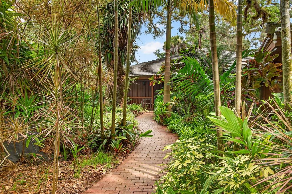 A brick path leads through a lush tropical garden toward a wooden house exterior — 3460/3480 RIVER RUN LANE, FORT MYERS, FL
