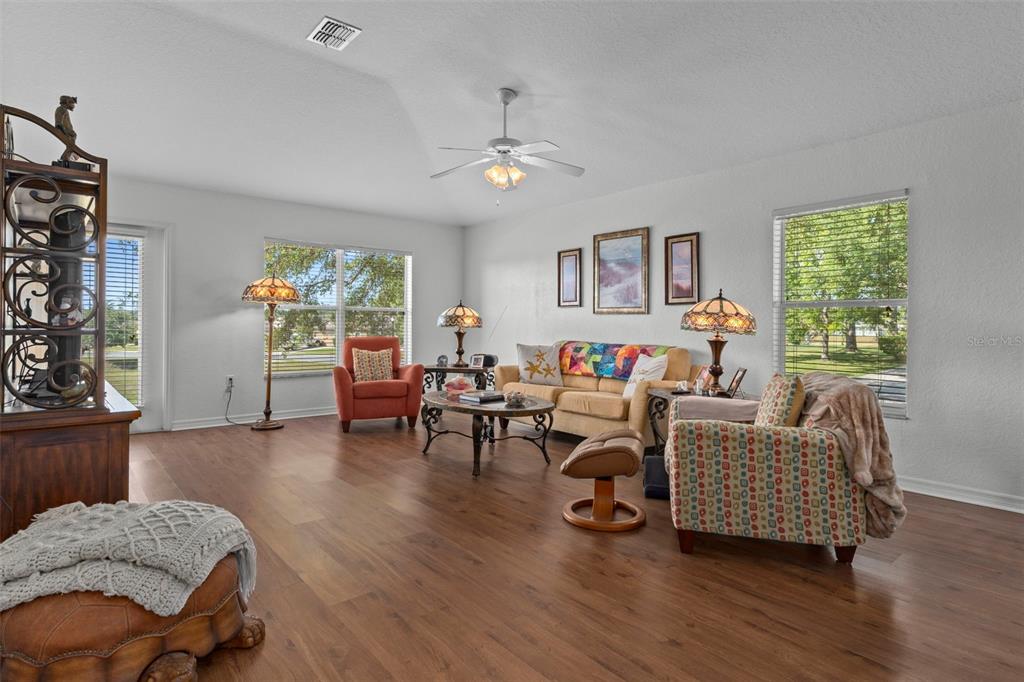 A living room features wood-look flooring, a ceiling fan, and multiple seating areas — 7228 CRESTED ORCHID DRIVE, BROOKSVILLE, FL