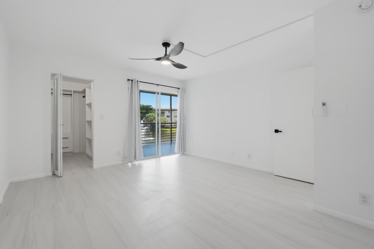 A bright, minimalist bedroom features white walls, light wood-look flooring, a ceiling fan, and balcony — 1805 Eleuthera Point # B2, Coconut Creek FL 33066, COCONUT CREEK, FL
