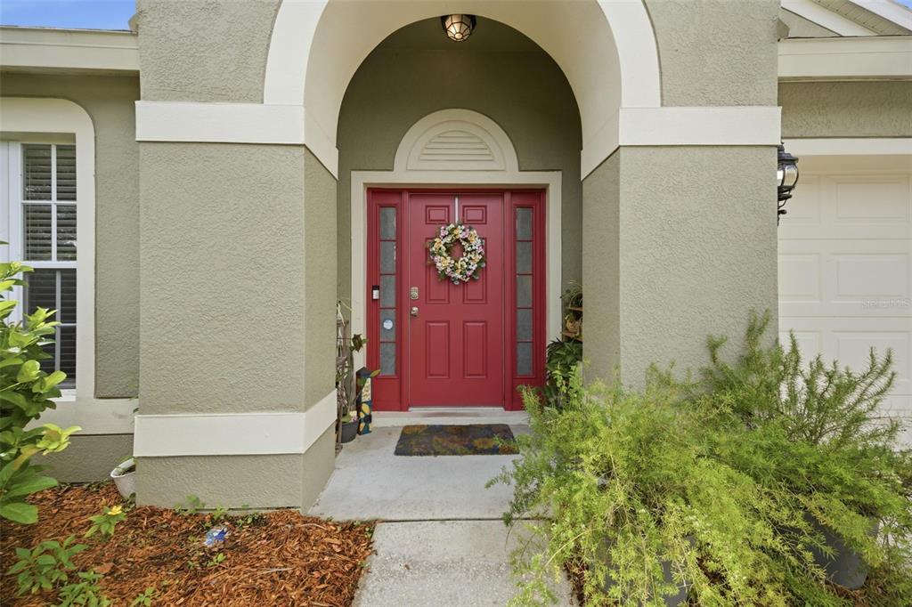 A red front door with sidelights is framed by a stucco archway and landscaping — 25114 LEXINGTON OAKS BOULEVARD, WESLEY CHAPEL, FL