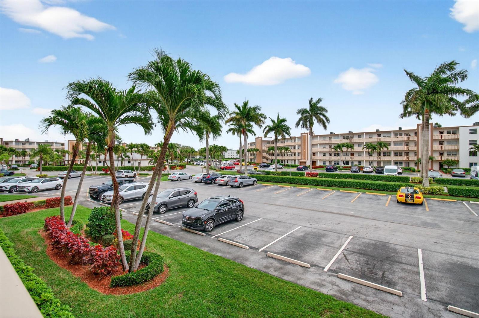 An outdoor parking lot with palm trees and multi-story apartment buildings under a blue sky — 2070 Guildford D, Boca Raton FL 33434, BOCA RATON, FL