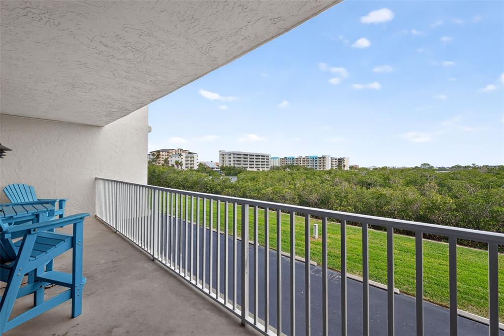 A balcony with blue chairs overlooks a grassy lawn, dense trees, and distant apartment buildings — 19451 GULF BOULEVARD UNIT 204, INDIAN SHORES, FL