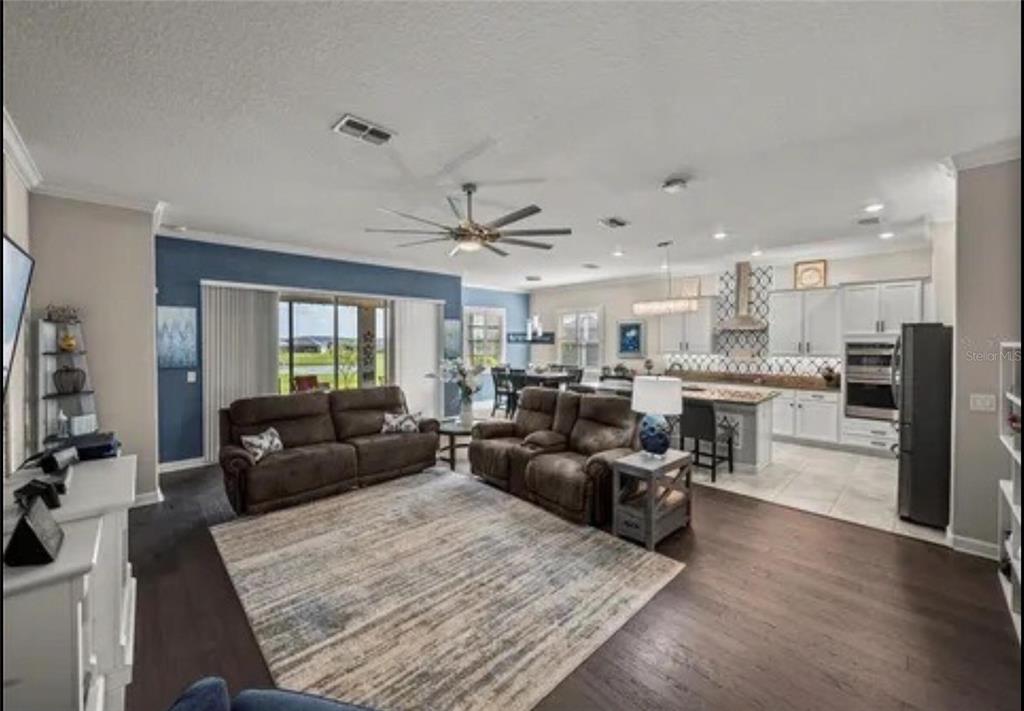 An open-concept living room with dark wood floors, brown sofas, and a kitchen in back — 31475 CABANA RYE AVENUE, SAN ANTONIO, FL