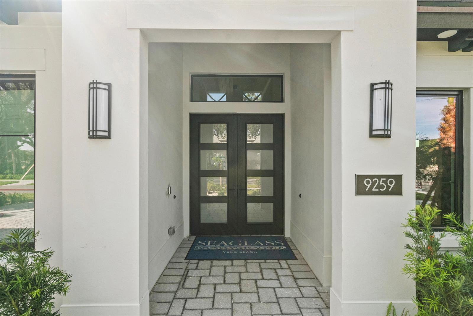 A white exterior entryway features a double door with glass panels and two wall sconces — 9259 Seaglass Court, Vero Beach FL 32963, VERO BEACH, FL