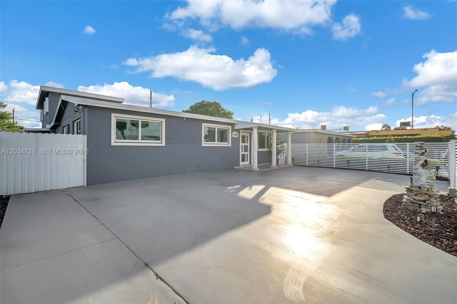 A gray single-story house features a large concrete driveway and a white picket fence — 380 W 30th St, Hialeah FL 33012, HIALEAH, FL