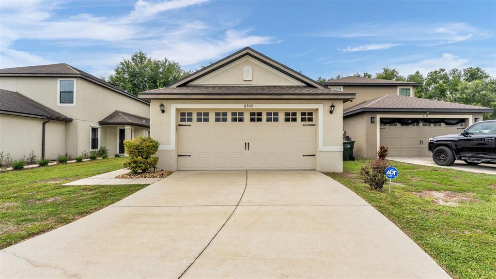 A beige house with a two-car garage and a concrete driveway sits under a blue sky — 6302 MANITOBA DRIVE, LAKELAND, FL