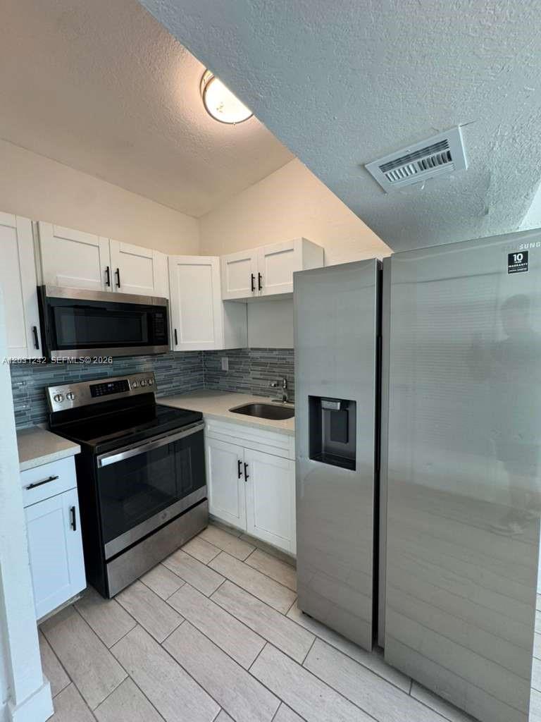 A kitchen features white cabinets, stainless steel appliances, a tiled backsplash, and light-colored flooring — RIVIERA BEACH, FL