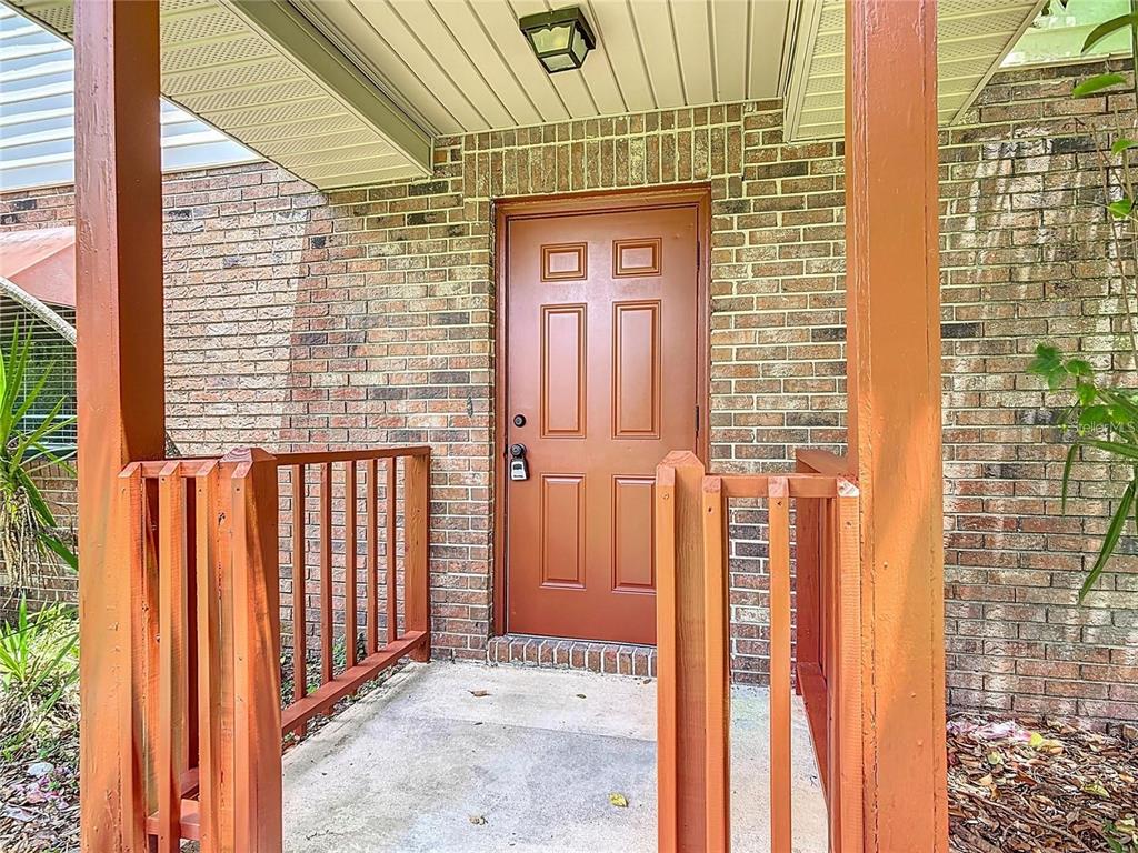 A brown front door is set within a brick exterior wall behind a wooden porch railing — 1591 REVERE LANE, DAYTONA BEACH, FL