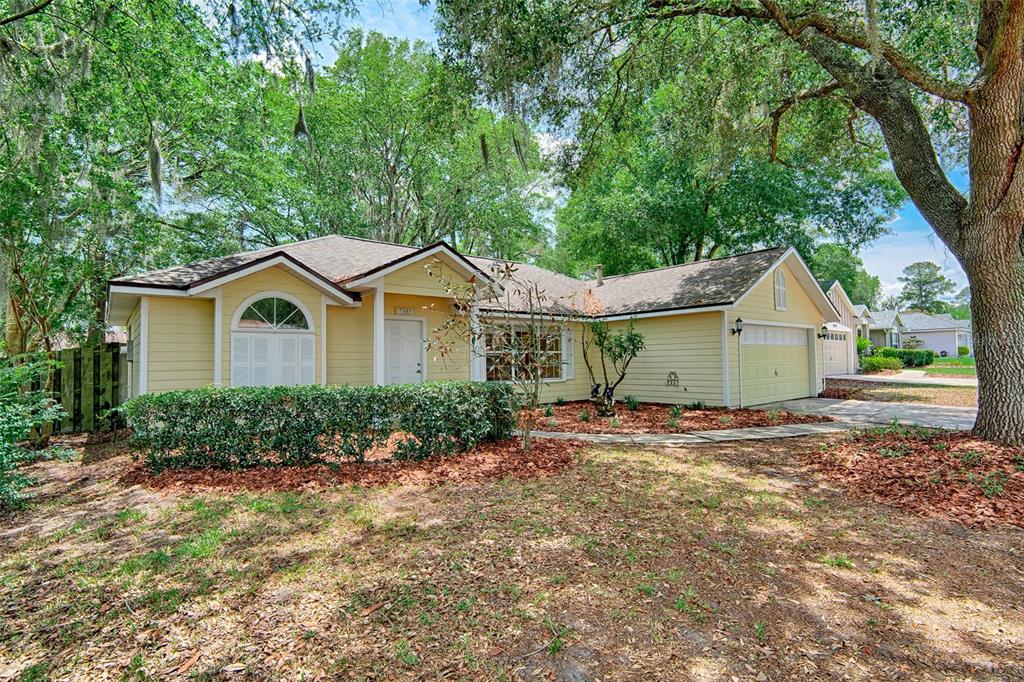 A yellow single-story house with a two-car garage sits under large trees — 7605 SW 51ST BOULEVARD, GAINESVILLE, FL
