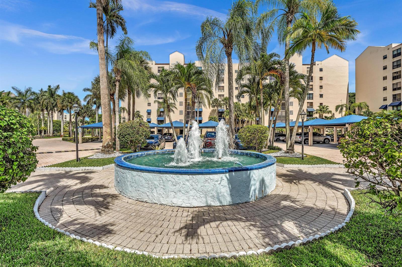 A circular fountain sits in the center of a paved courtyard surrounded by palm trees — 7564 Regency Lake Drive # 202, Boca Raton FL 33433, BOCA RATON, FL