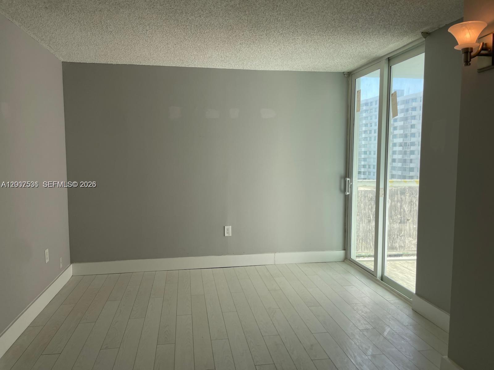 An empty room features gray walls, light wood-look flooring, a popcorn ceiling, and a sliding glass door — 5880 Collins Ave # 1006, Miami Beach FL 33140, MIAMI BEACH, FL