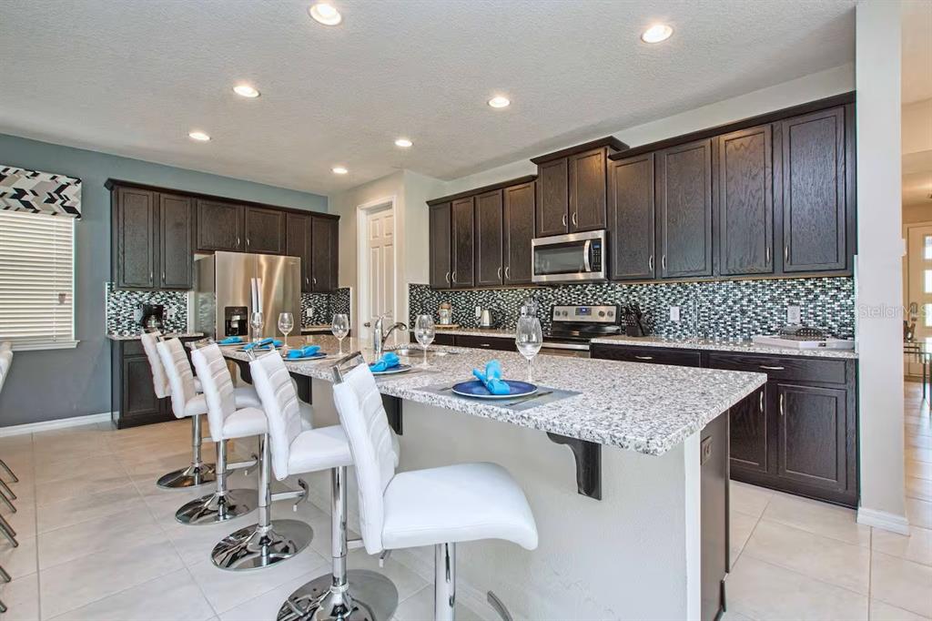 A kitchen features dark wood cabinets, granite countertops, a tiled backsplash, and white bar stools — 9015 SUNSHINE RIDGE LOOP, KISSIMMEE, FL