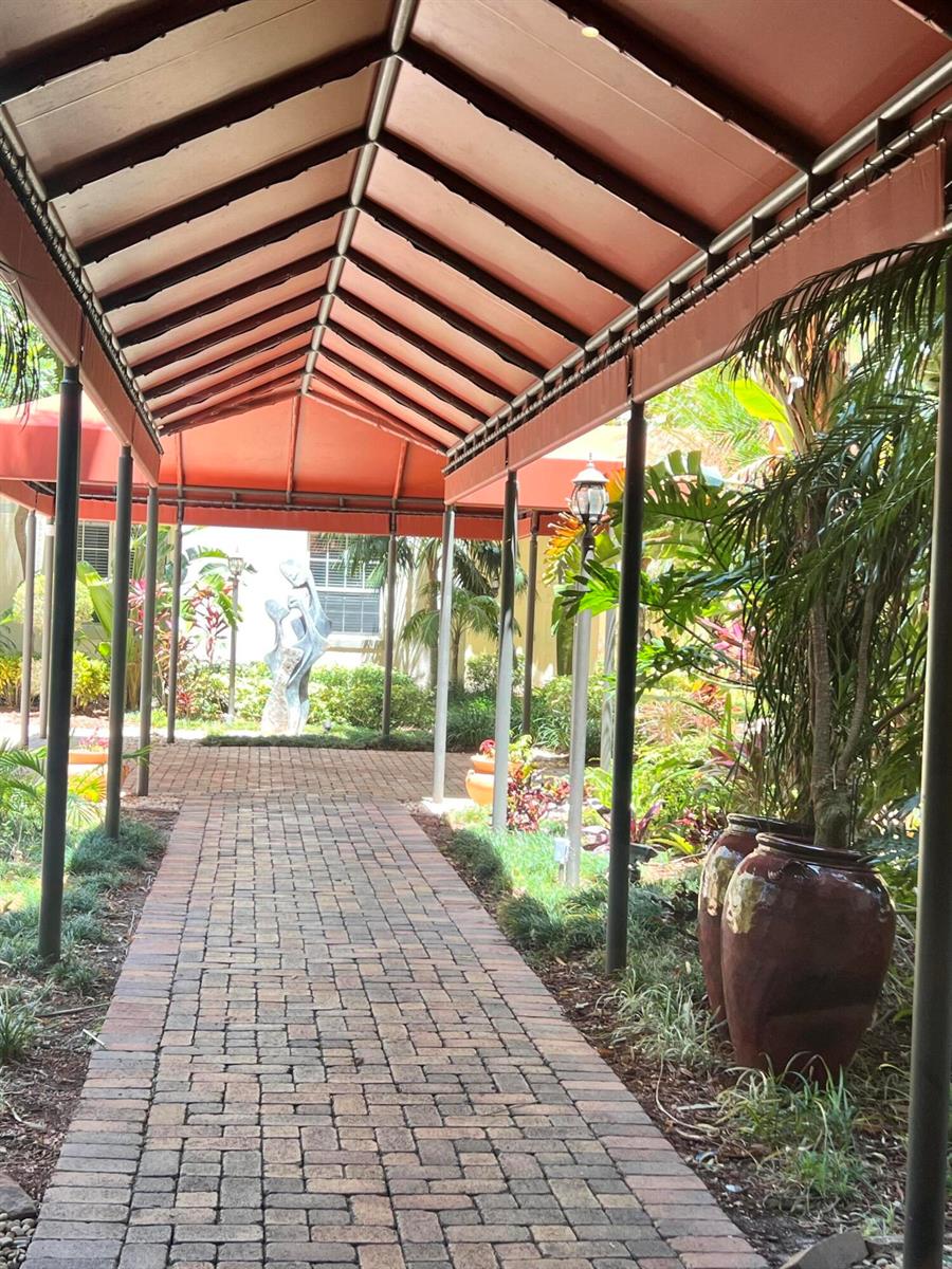 A brick walkway leads under a covered orange awning toward a sculpture in a garden — 651 Okeechobee Boulevard # 706, West Palm Beach FL 33401, WEST PALM BEACH, FL