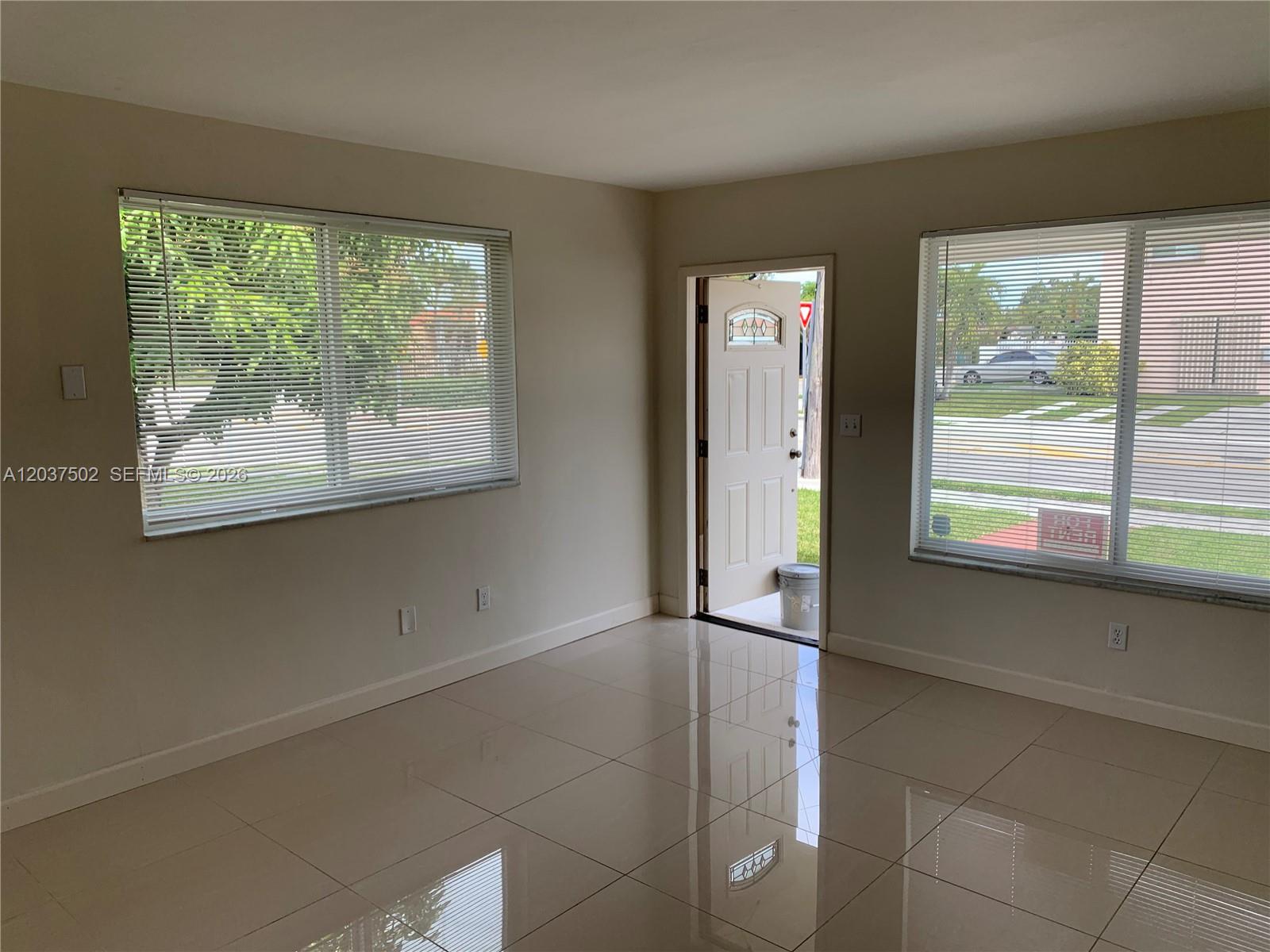 A bright, empty room features light-colored tile flooring, two large windows with blinds, and an open door — 1200 SW 31st Ave, Miami FL 33135, MIAMI, FL