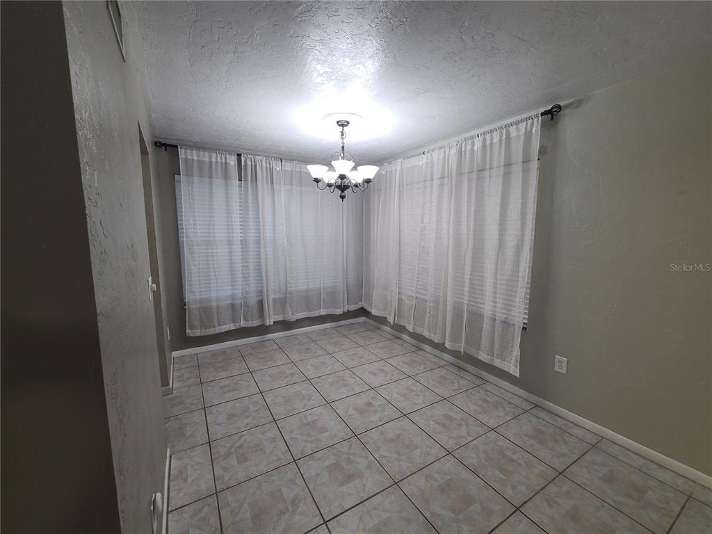 An empty dining room features tile flooring, a central chandelier, and white curtains over windows — 18101 SANDALWOOD DRIVE, WILDWOOD, FL