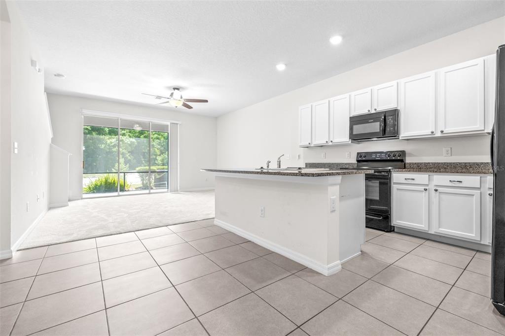A kitchen with white cabinets, granite countertops, and an island opens to a carpeted living area — 32960 KALOKO ROAD, WESLEY CHAPEL, FL