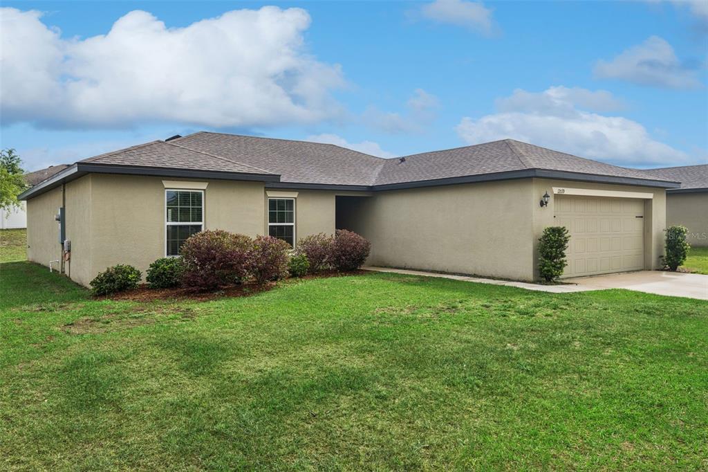 A single-story tan stucco house with a two-car garage and a grassy front yard — 12019 INDIAN GRASS WAY, LEESBURG, FL