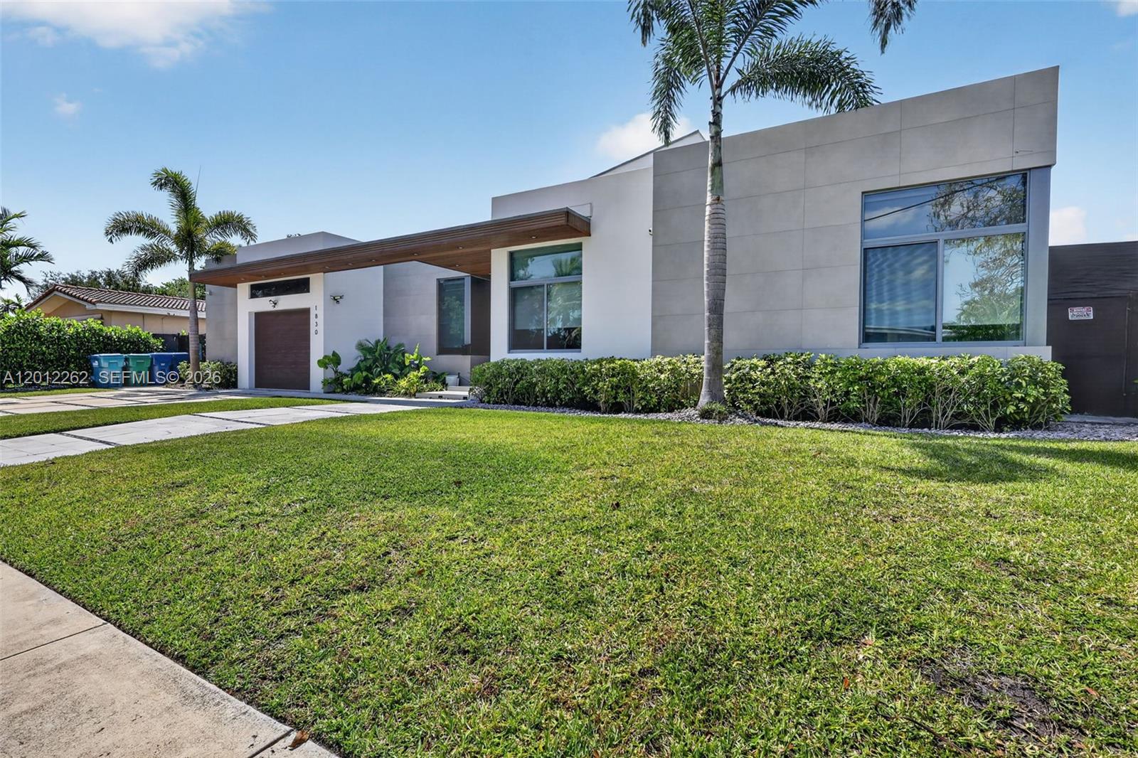 A modern, single-story house with a flat roof, wood-accented entryway, and a manicured front lawn — 1830 NE 193rd St, Miami FL 33179, MIAMI, FL