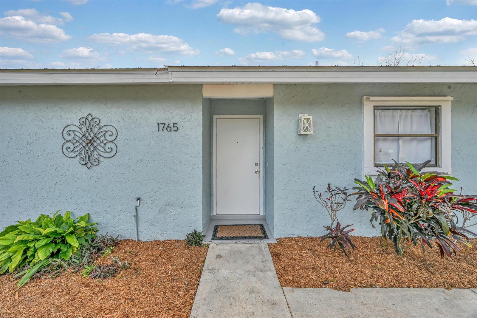 The exterior of a light blue house features a white front door and landscaping — 1765 24th Place SW, Vero Beach FL 32962, VERO BEACH, FL