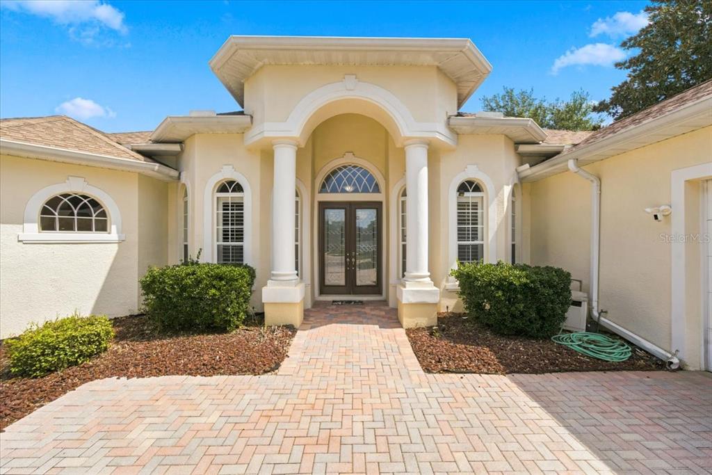 The exterior of a light-colored house features a brick walkway leading to a pillared entryway — 13519 BARKINGSIDE PLACE, SPRING HILL, FL