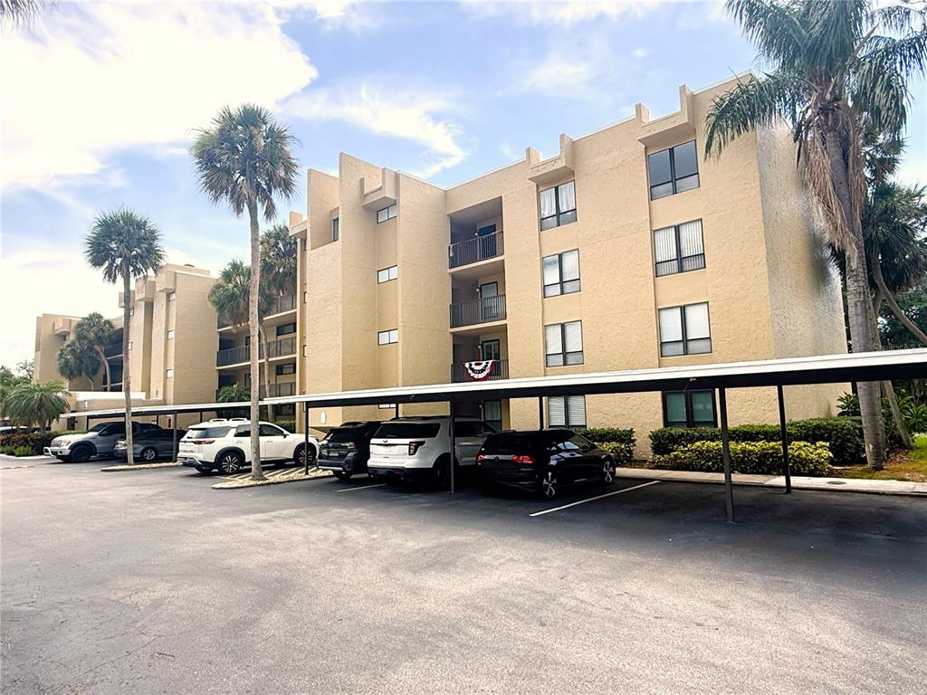 A multi-story tan apartment building features covered parking spaces and several palm trees — 2614 COVE CAY DRIVE UNIT 204, CLEARWATER, FL