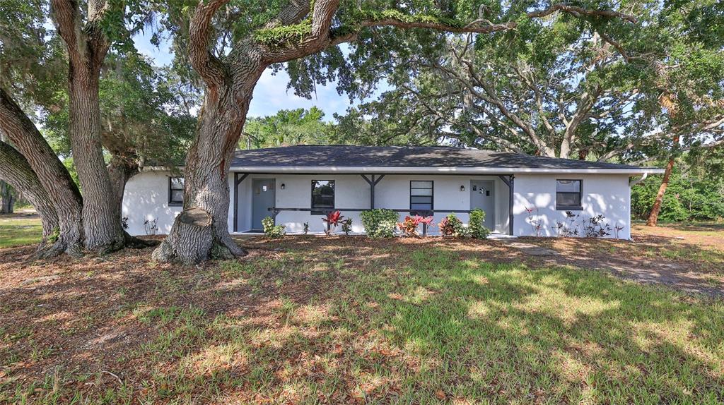 A single-story duplex with a covered porch sits under large oak trees on a grassy lot — 1802 N FERN PALM DRIVE, EDGEWATER, FL