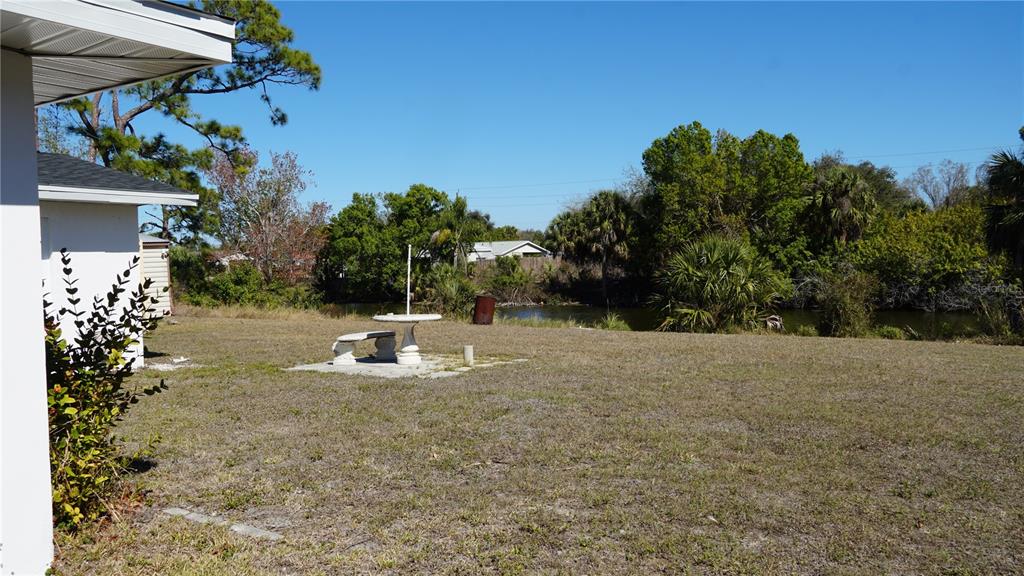 A grassy backyard features a stone table and bench set overlooking a small pond — 3552 MIDDLETOWN STREET, PORT CHARLOTTE, FL