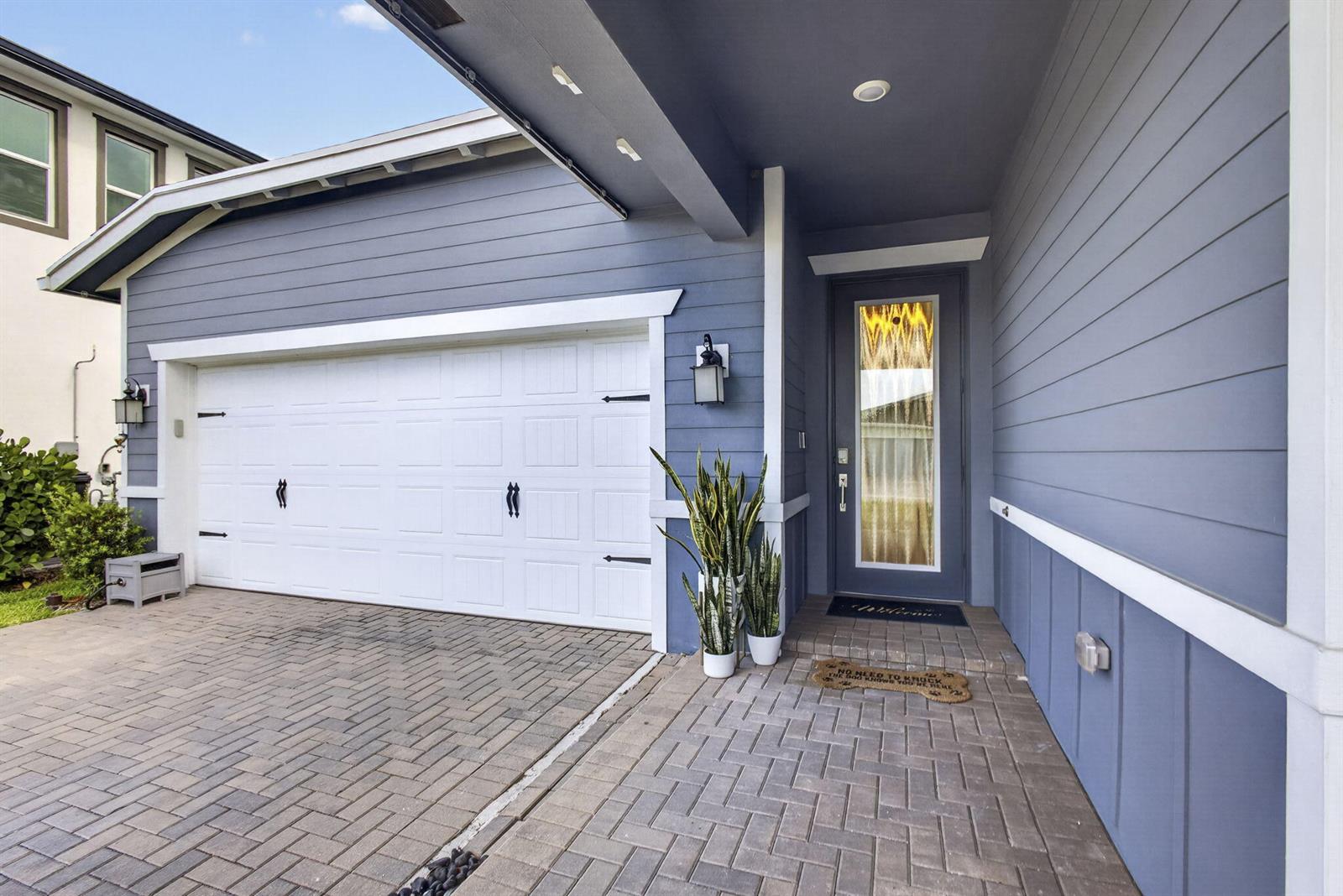 A blue house exterior features a white garage door, a front door, and brick pavers — 1919 Wandering Willow Way, FL 33470, FL