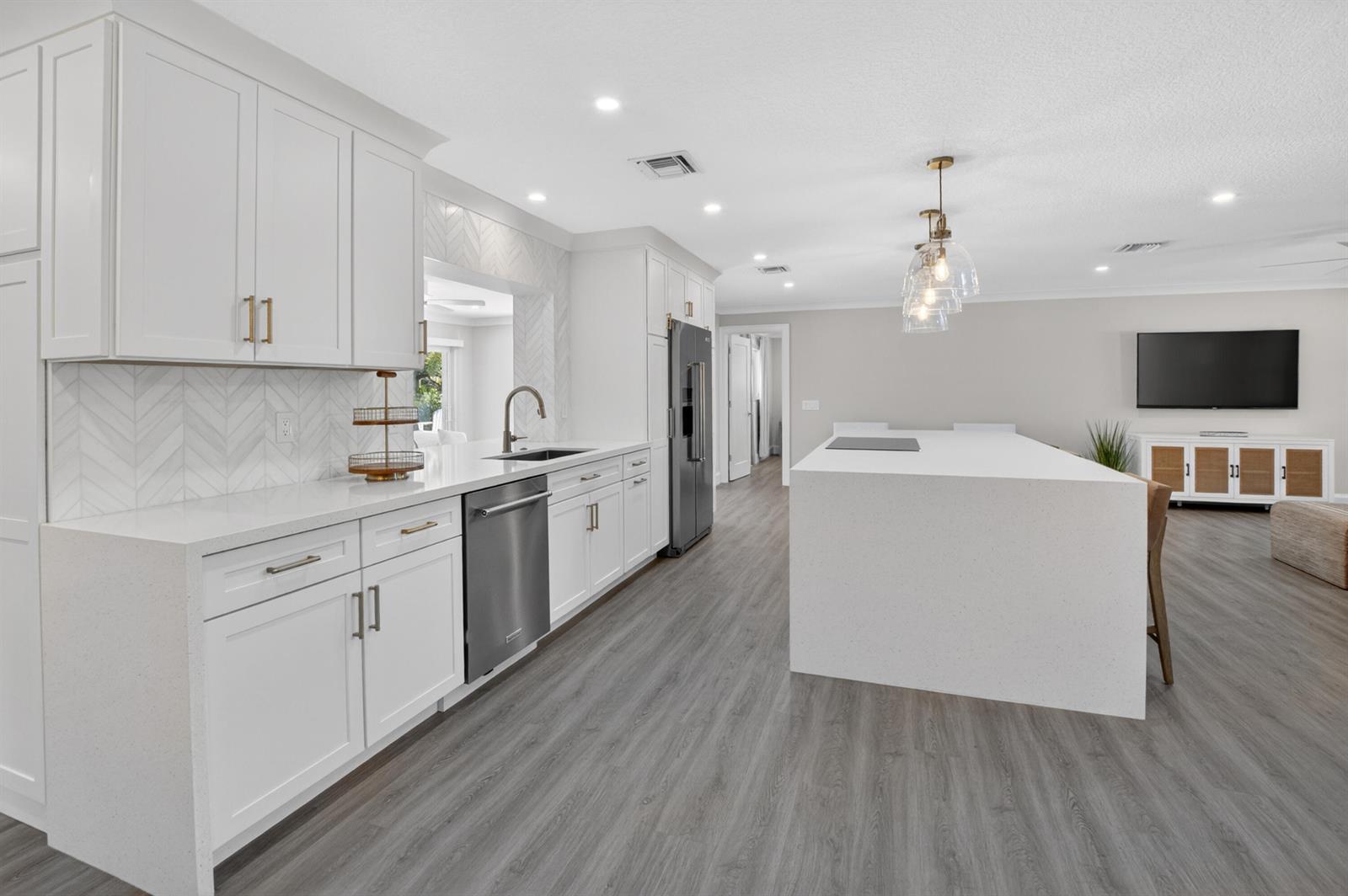 A modern kitchen features white cabinetry, a large quartz island, and gray wood-look flooring — 135 SE 28th Court, Boynton Beach FL 33435, BOYNTON BEACH, FL