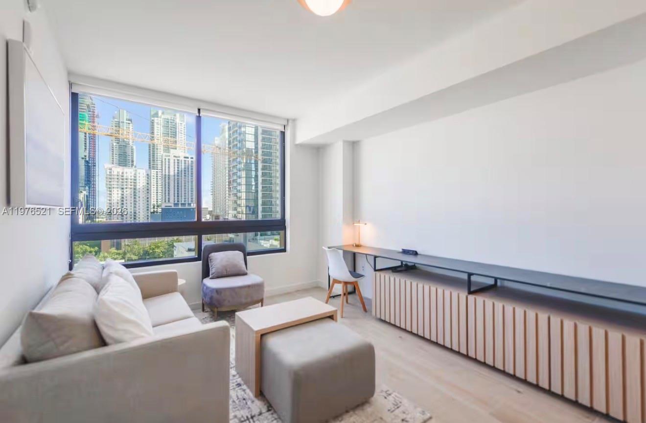 A modern living room features a sofa, a desk, and large windows overlooking city skyscrapers — 225 N Miami Ave # 1103, Miami FL 33128, MIAMI, FL