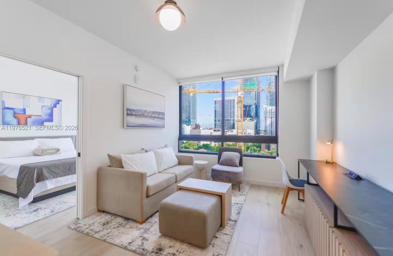 A living room with a sofa, desk, and large window overlooking a city construction site — 225 N Miami Ave # 1103, Miami FL 33128, MIAMI, FL
