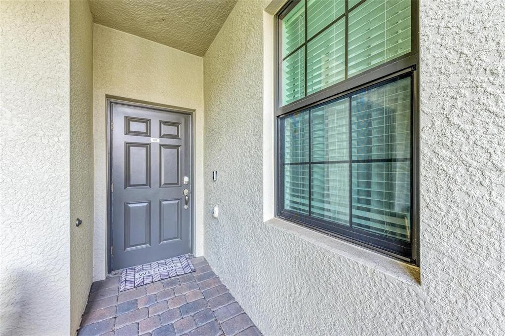 A gray front door with a welcome mat sits on a paved entryway beside a window — 5685 PALMER CIRCLE UNIT 105, BRADENTON, FL