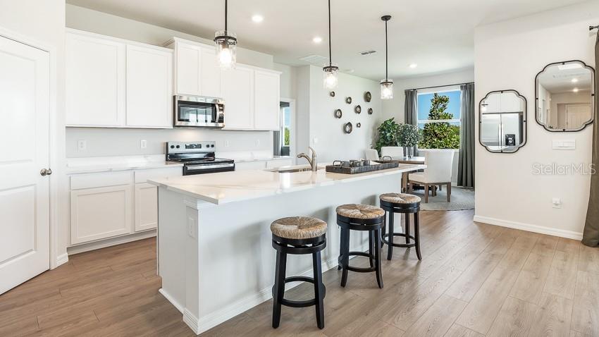 A modern kitchen features white cabinets, a quartz-topped island with stools, and light wood flooring — 10433 WOODLAND GARDEN WAY, PARRISH, FL