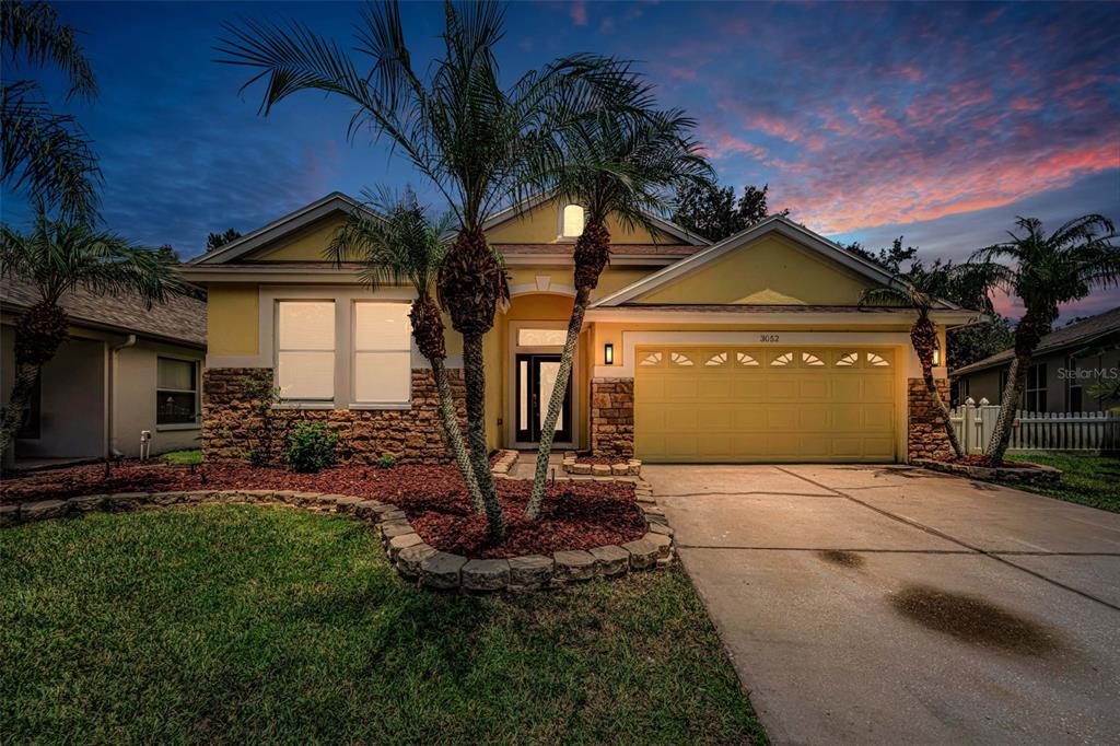 A single-story house with a yellow exterior, stone accents, and a two-car garage at dusk — 3052 SUNWATCH DRIVE, WESLEY CHAPEL, FL