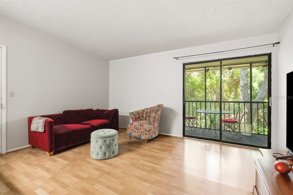 A living room with light wood floors, a red sofa, patterned chair, and sliding glass doors — 3295 FOX CHASE CIRCLE N UNIT 207, PALM HARBOR, FL