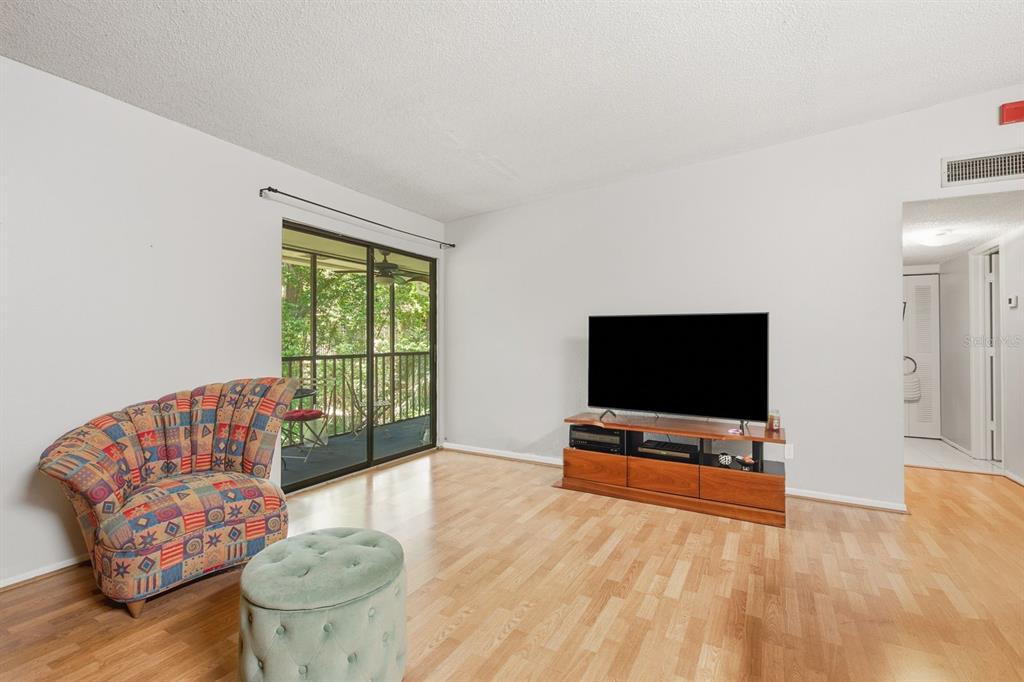 A living room features light wood flooring, a patterned armchair, a television, and a balcony — 3295 FOX CHASE CIRCLE N UNIT 207, PALM HARBOR, FL