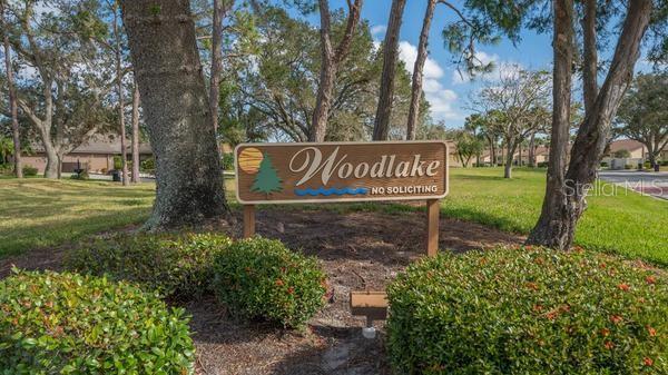 A wooden sign for Woodlake stands in a grassy area surrounded by trees and bushes — 5905 DORAL DRIVE, SARASOTA, FL