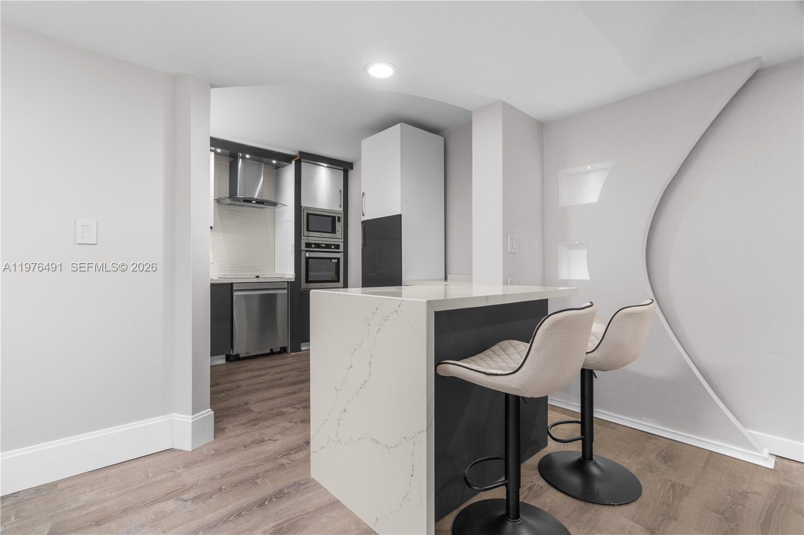 A modern kitchen features a white quartz island with two bar stools and dark cabinetry — 3730 Inverrary Dr # 2H, Lauderhill FL 33319, LAUDERHILL, FL