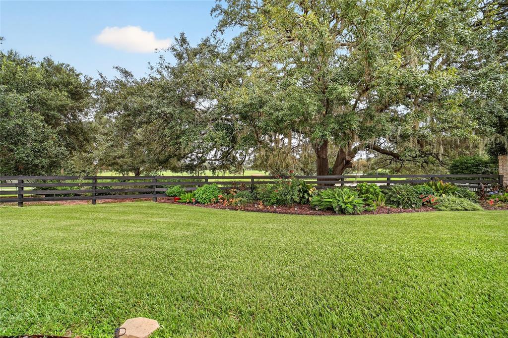A grassy backyard features a dark wooden fence, mature trees, and a landscaped garden bed — 8293 SE 176TH LAWSON LOOP, THE VILLAGES, FL