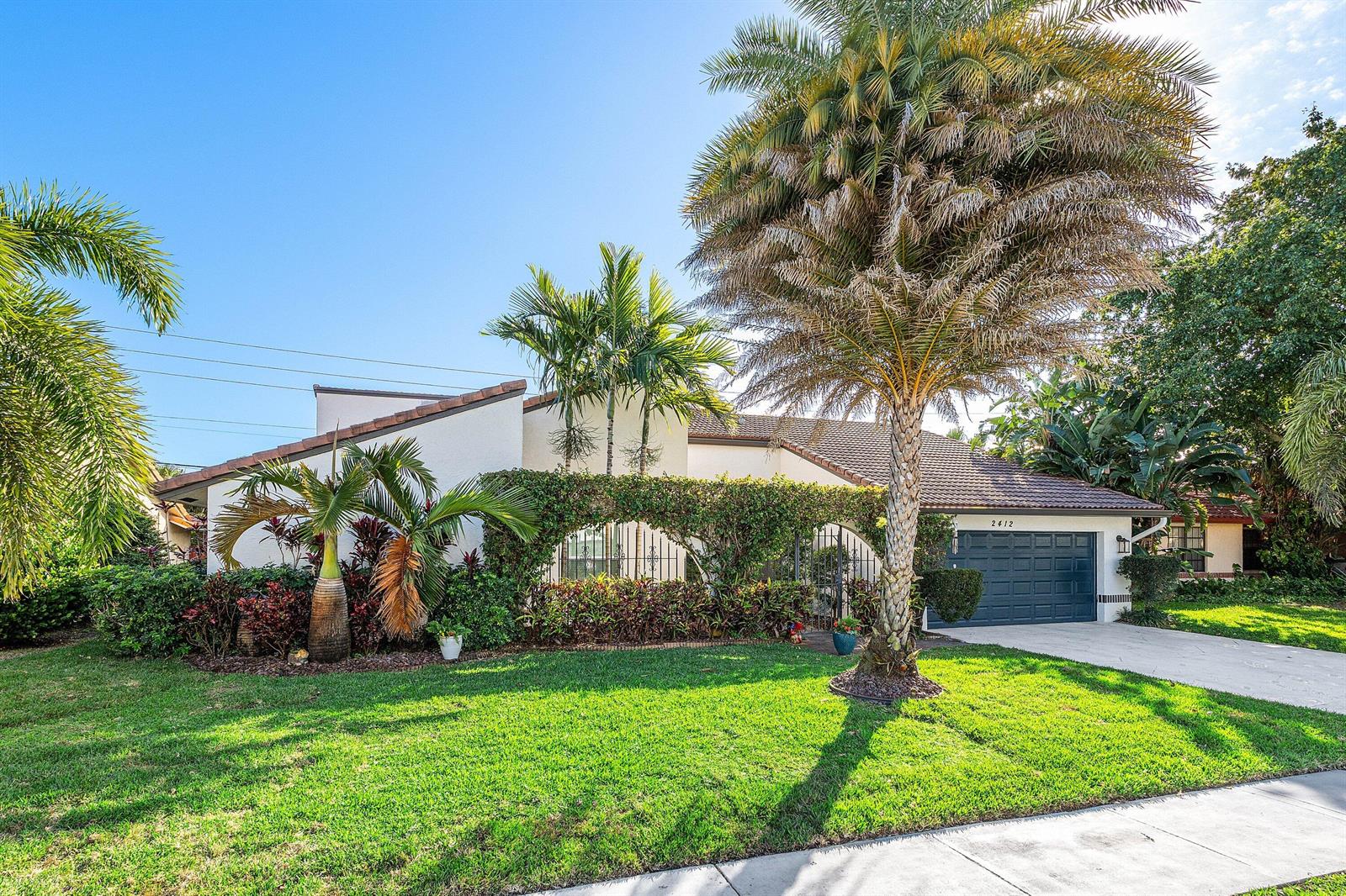 A single-story house with a dark garage door, palm trees, and a vine-covered front wall — 2412 SW 23rd Cranbrook Drive, Boynton Beach FL 33436, BOYNTON BEACH, FL