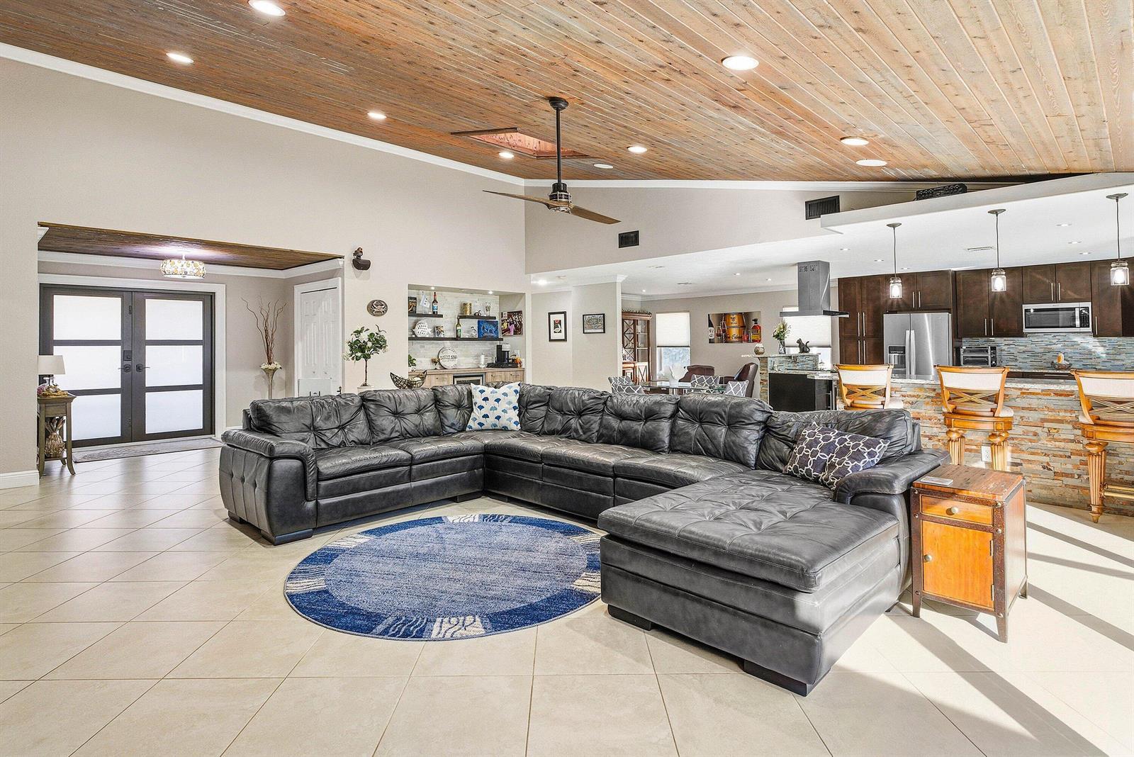 A large open-concept living room features a dark leather sectional, wood-plank ceiling, and kitchen island — 2412 SW 23rd Cranbrook Drive, Boynton Beach FL 33436, BOYNTON BEACH, FL