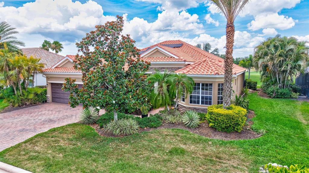 A single-story house with a tiled roof, brick driveway, and lush landscaping under a cloudy sky — 7511 HERITAGE GRAND PLACE, BRADENTON, FL