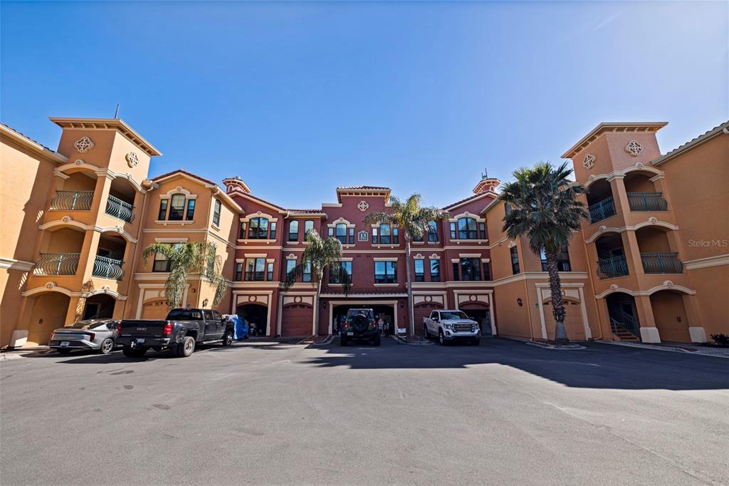 A multi-story apartment building with stucco walls, arched balconies, and a paved parking area — 2773 VIA CIPRIANI UNIT 1321B, CLEARWATER, FL