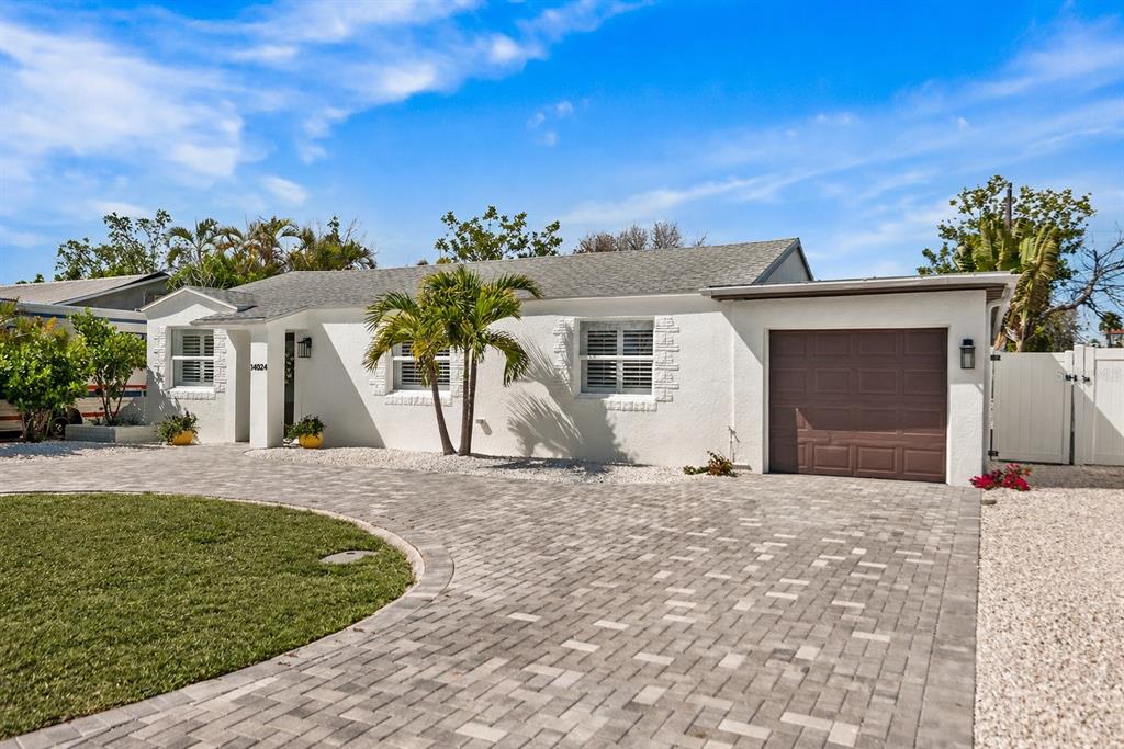 A white single-story house features a paver driveway, a brown garage door, and palm trees — 14024 N BAYSHORE DRIVE, MADEIRA BEACH, FL