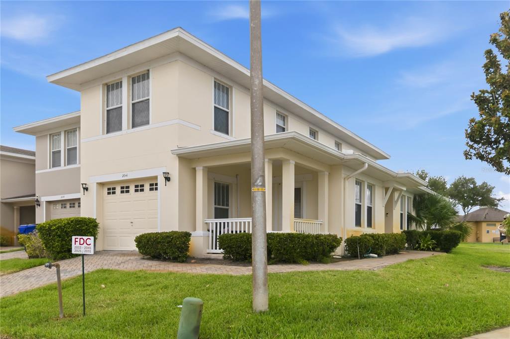 A two-story beige house features a front-facing garage, a covered porch, and landscaped bushes — 2041 LACIE JO LANE, KISSIMMEE, FL