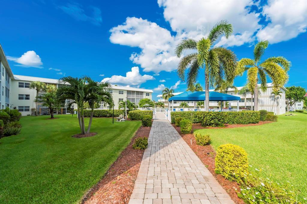 A paved walkway leads through a grassy courtyard toward a blue-canopied pool area with palms — 9826 Marina Boulevard # 1015, Boca Raton FL 33428, BOCA RATON, FL