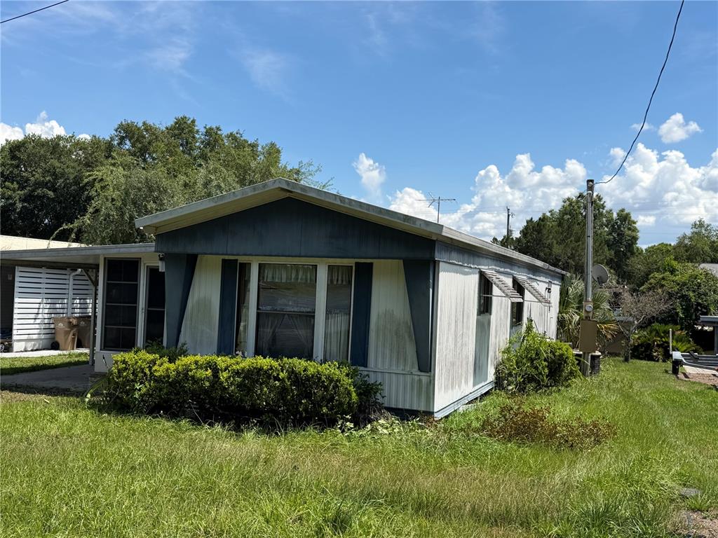 A single-story manufactured home with a carport and overgrown lawn under a blue sky — 13440 PALM DRIVE UNIT 7, ASTATULA, FL